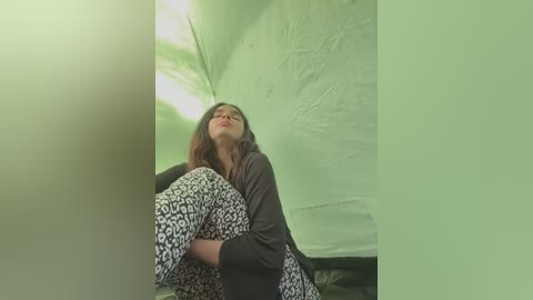 Media: A video of a young woman with light brown skin, wearing a dark long-sleeve top and leopard print leggings, sitting inside a green tent with a white canopy.