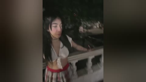 Media: A dimly lit video of a young woman with long dark hair, wearing a white crop top and a red skirt, standing on a balcony with green foliage in the background.