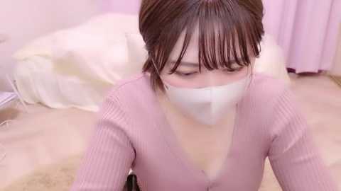 Video of an East Asian woman with straight brown hair, wearing a pink ribbed sweater and a white face mask, kneeling on a beige carpeted floor in a softly lit room with pastel-colored curtains and a white bed.