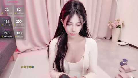 Media: A video of an Asian woman with long black hair, wearing a white top, sitting indoors with a phone, surrounded by pink curtains and a vase of flowers.