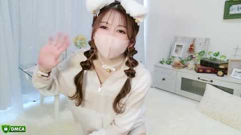 Media: Video of a young Asian woman in a white cat costume, waving with a face mask on, in a bright, minimalist room with white furniture and decor.