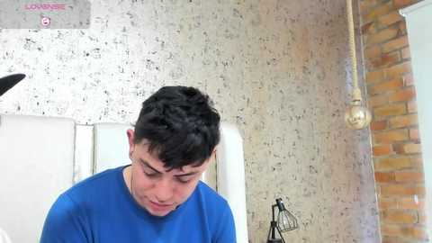 Media: Video of a young man with short dark hair, wearing a blue shirt, looking down in a room with textured wallpaper and exposed brick wall.