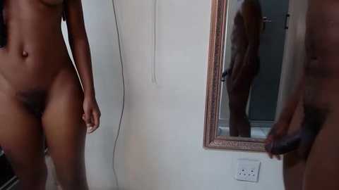 Media: Video of a naked Black woman standing in front of a mirror, holding a dark dildo, in a minimalist bathroom with white walls and a silver door.