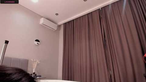 Media: A video of a modern, minimalist bathroom with beige walls, a large window covered by brown curtains, a white air conditioner, and a small white clock on the left.