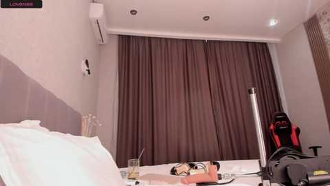 Media: Video of a modern hotel room with white walls, brown curtains, white bed, air conditioning unit, desk with laptop, and gaming chair in the background.