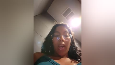 Media: Video of a young woman with curly black hair, wearing glasses and a teal top, captured from a low angle, in a dimly lit room with a white ceiling and a bright light source above.