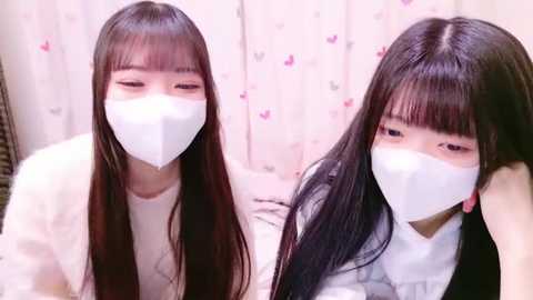 Media: Video of two East Asian women with long black hair, wearing white masks, white tops, and white backgrounds adorned with pink heart patterns.