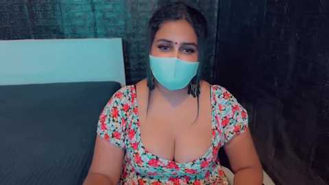 Media: A video of a South Asian woman with dark hair, fair skin, and a large bust, wearing a floral dress, face mask, and traditional bindi, sitting on a bed with a dark headboard in a dimly lit room.