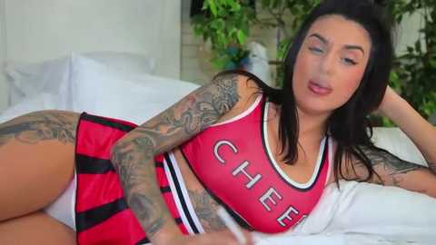 Media: Video of a tattooed woman with dark hair, wearing a red cheerleader outfit, lying on a bed in a bright, indoor setting.