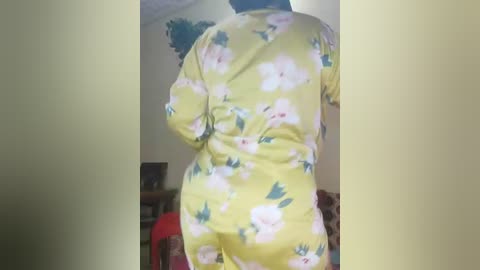 Media: Video of a person wearing a yellow pajama set with white and pink flower patterns, standing in a dimly lit room with a shelf and a red chair in the background.