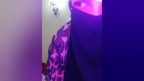 Media: A video of a person with a dark mask covering the lower face, wearing a camouflage-patterned shirt. The background is dimly lit with purple and blue lighting, creating a mysterious and somewhat eerie atmosphere.