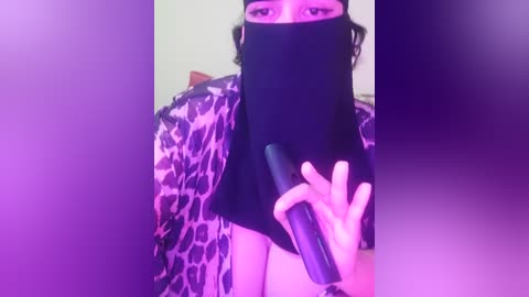 Media: A video of a person wearing a black mask, leopard-print robe, and holding a black vibrator. The background is blurred, featuring purple lighting.