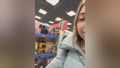 Media: Video of a young woman with straight, shoulder-length blonde hair, partially obscured, in a store with shelves full of colorful toys and a grid-patterned ceiling.