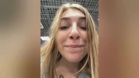 Media: Video of a young, fair-skinned, blonde woman with wet hair, wearing a light-colored top, smiling with closed eyes in a tiled bathroom with a grid-patterned ceiling.