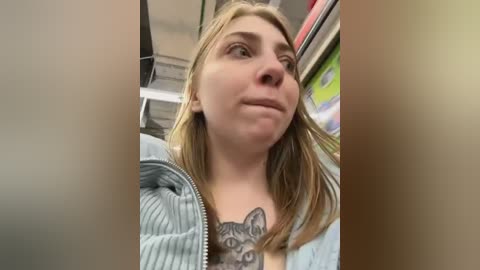 Media: Video of a young woman with fair skin, blonde hair, and a grey hoodie, showing a large cat tattoo on her chest. She stands in a brightly lit store, with colorful posters in the background.