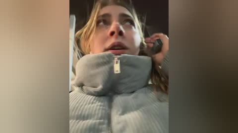 Media: Video of a young woman with light skin, wearing a light grey, ribbed zip-up hoodie, talking on a phone, her expression serious.