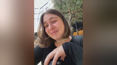 Media: Video of a young woman with shoulder-length brown hair, wearing a black top and a choker necklace, smiling while holding a camera, outdoors with a modern building and tree in the background.