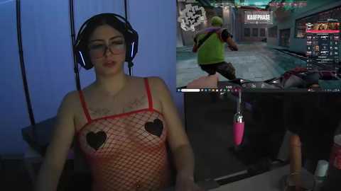 Media: Video of a fair-skinned woman with glasses and dark hair, wearing a red fishnet top with black heart cutouts, seated. Scene split into two: top shows her in a gaming setup, bottom shows a virtual reality game.