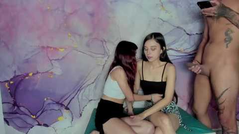 Media: Video: Two young women, one in a white crop top, black shorts, the other in a black crop top, black skirt, sitting on a table. Background: Abstract, colorful painting.