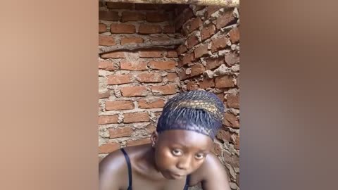 Media: Video of a young African girl with dark skin, wearing a black braided hairstyle and a black tank top, crouching in a rustic, red brick room with a worn, earthy background.