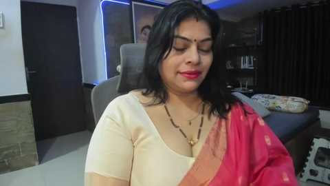 Media: Video of a smiling South Asian woman with dark hair, wearing a white blouse and pink saree, seated in a modern, dimly lit room with a black chair and shelves.