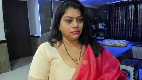 Media: Video of a middle-aged woman with long black hair, wearing a red saree and gold necklace, sitting indoors with a serene expression.