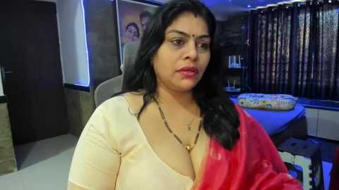 Media: Video of a South Asian woman with long black hair, wearing a red saree, seated indoors, with a beige blouse and gold necklace. Background features a modern, dimly lit room with a large window and bookshelves.