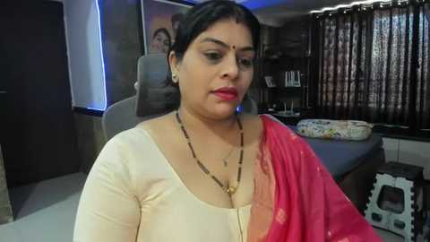 Media: Video of a South Asian woman in a cream blouse, wearing a red sari with gold embroidery, and a gold necklace, seated in a modern office setting.