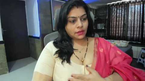 Media: Video of an Indian woman with long black hair, wearing a pink and beige sari, seated indoors with a neutral expression.