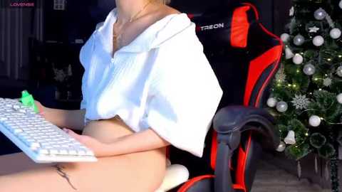 Media: Video of a blonde woman in a white, short-sleeved blouse, wearing nothing below the waist, sitting in a red and black gaming chair, typing on a keyboard.