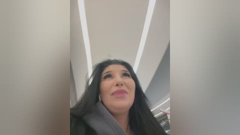 Media: Video of a woman with long black hair, wearing a grey coat, looking upward with a neutral expression, standing in a modern, white-walled interior with recessed lighting.