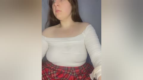 Media: A video of a young Caucasian woman with long brown hair, wearing a white off-the-shoulder top and a red plaid skirt, looking pensive against a plain blue background.