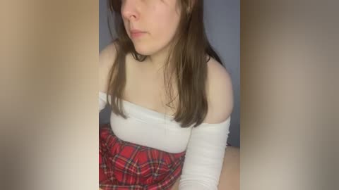 Media: Video of a young woman with fair skin and brown hair, wearing a white off-shoulder top and red plaid skirt, in a dimly lit room.