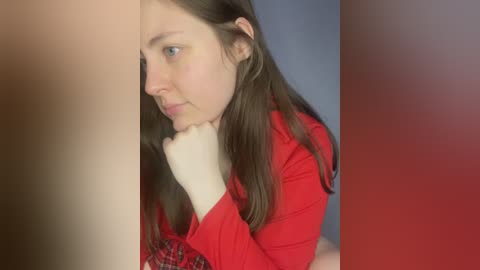 Media: Video of a young woman with light skin and straight brown hair, wearing a red shirt, resting her chin on her hand, looking pensive. Background is a blurry gradient of red and blue.