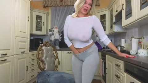 Media: Video of a blonde Caucasian woman with large breasts in a tight white crop top and form-fitting light grey leggings, standing in a modern kitchen with cream-colored cabinets.