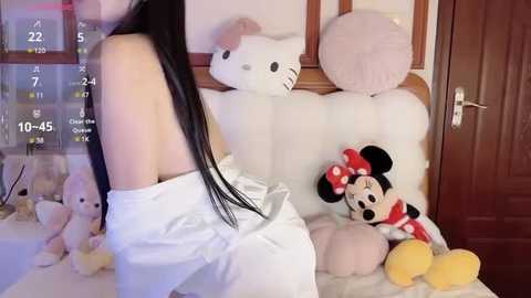 Media: Video of a woman with long black hair wearing a white robe, sitting on a bed with plush toys including Mickey Mouse and Minnie Mouse, in a cozy room with a wooden door.