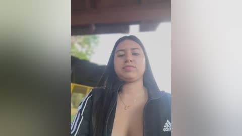 Media: Video of a Latina woman with long black hair, fair skin, wearing a black Adidas jacket with a V-neck, standing outdoors in a blurred background.