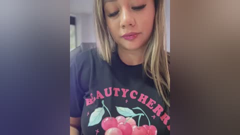 Video of a young woman with light brown hair, wearing a black t-shirt with \"Beauty Cherry\" logo, standing indoors, eyes closed, slightly smiling.