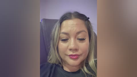 Video of a young Asian woman with straight, shoulder-length blonde hair, wearing black clothing, sitting in a dimly lit room. She has makeup on, with long eyelashes and pink lipstick, appearing calm and composed.