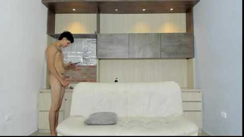 Video of a nude man with a small penis, standing in a minimalist, modern room with white walls, a white couch, and wooden cabinets.