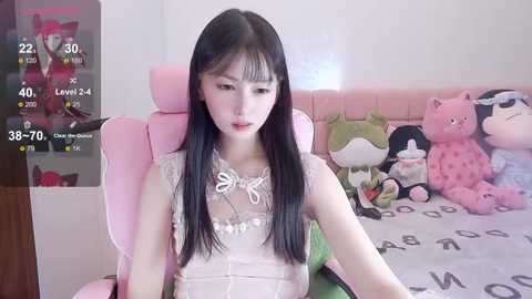 Media: Video of an East Asian woman with long black hair, wearing a sheer, lace-trimmed top, seated in a pink gaming chair, surrounded by plush toys in a cozy, pastel-themed room.