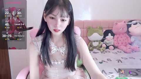 Media: Video of a young East Asian woman with long black hair, wearing a white lace dress, sitting on a pink chair in a room with stuffed animals.