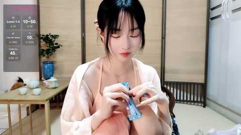 Media: A video of an Asian woman with fair skin, wearing a light robe, carefully unwrapping a condom packet indoors. Background includes a table with teacups and a plant.