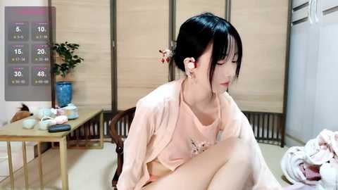 Media: A video of a young Asian woman with fair skin, black hair, and bangs, wearing headphones and a pale pink top, sitting on a wooden chair in a minimalist room with a wooden screen backdrop.