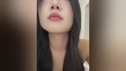 Media: Video of a young Asian woman with fair skin, long black hair, and full lips. She's wearing a sleeveless top. The background is blurred, featuring a beige wall and a hint of a bed.