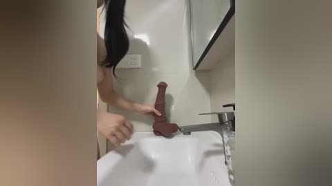 Media: A video of a woman with long black hair, nude, holding a large, phallic-shaped object against a white bathroom sink.