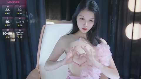 Media: Video of an East Asian woman with pale skin and long black hair, wearing a pink feathered top, forming a heart shape with her hands, sitting on a white chair in a dimly lit room with dark curtains.