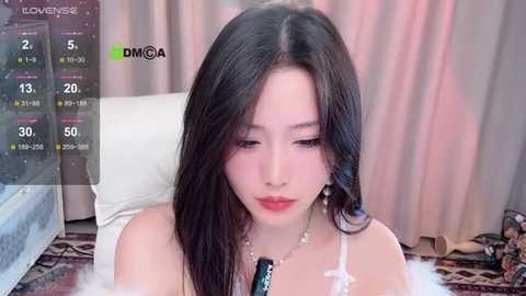 Media: Video of an East Asian woman with long, straight black hair and fair skin, wearing a white lace bra, sitting on a bed with beige curtains and a blue blanket.