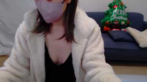 Media: Video of a woman in a white fuzzy robe, black top, pink face mask, sitting on a blue couch, next to a green Christmas bag.