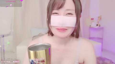 Media: A young Asian woman with light skin and short brown hair, wearing a white face mask and a spaghetti strap top, holding a metallic cup, smiling in a softly lit room.